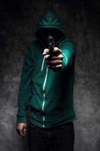 An image depicting a young adult student aiming a handgun at the camera and standing in front of a concrete wall, dark dramatic lighting spotlighting the weapon. His face is obscured by a hooded sweatshirt. Representing gun violence and school shootings or universities, etc. Vertical image.